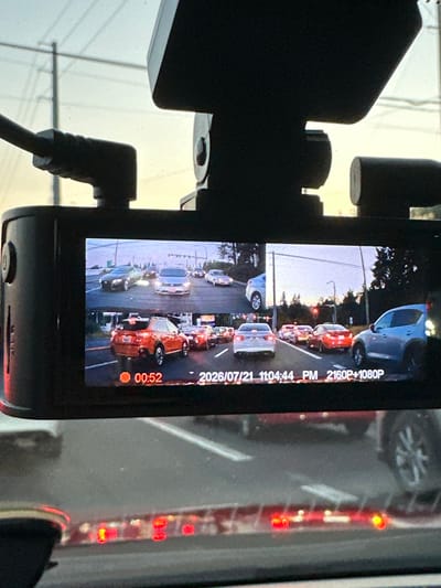 The image is a dashcam view of traffic on a street in North Lynnwood, United States. The dashcam is mounted on the windshield of a vehicle. The screen displays two simultaneous feeds of the road ahead. The top feed shows a lane of traffic with several cars. The bottom feed shows another lane of traffic with more cars, including a red SUV in the foreground. The time stamp on the screen reads "00:52 2026/07/21 11:04:44 PM". The sky is overcast, suggesting it might be evening or a cloudy day. The environment appears to be suburban or urban with power lines and trees visible in the background. The scene depicts a traffic jam or slow-moving traffic.