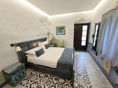 The image depicts a bedroom in Pozzuoli, Italy. The room features a large bed with white linens and dark blue accent pillows and a throw blanket. The bed frame and a matching bedside table are painted a distressed teal color, adding a rustic charm. The walls are adorned with a nature-themed mural, featuring trees, foliage, and a small painting of a bird on water. A wall-mounted lamp with a white shade is positioned above the bedside table.
The floor is covered with an intricate black and white patterned tile, interspersed with areas of speckled terrazzo. To the right of the bed, there is a dark wooden door with glass panels, suggesting an entrance to another part of the residence or an exterior exit. Further down the hallway, another similar door is visible, along with light gray curtains hanging by a window. The room is illuminated by recessed ceiling lights, creating a bright and airy atmosphere. The overall aesthetic is a blend of rustic, artistic, and Mediterranean styles. There is no visible text in the image. The time of day is not discernible, and the weather is irrelevant to the indoor scene.