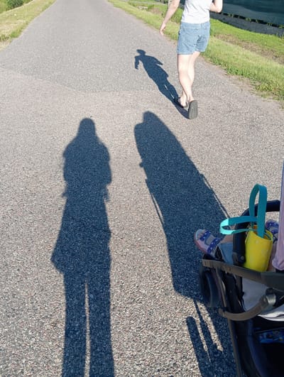 The image depicts a paved path, likely a sidewalk or bike lane, in Dybów Kolonia, Poland. The path is bordered by grass and vegetation on the left and a fence and more grass on the right. The sun is casting long shadows, indicating it is either morning or late afternoon. In the foreground, a stroller is visible with a child's legs and feet, wearing sandals, extending out. A yellow bucket with a teal handle is attached to the stroller. In the background, a person in denim shorts and a t-shirt is walking away from the camera, their shadow cast on the path. Further back, two other distinct shadows of people are also visible on the pavement, suggesting a family outing. The overall scene conveys a sense of leisure activity in an outdoor, possibly suburban or rural, setting within Dybów Kolonia.