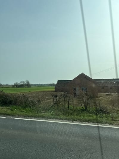 The image depicts a rural scene in Thorpe Waterville, United Kingdom, viewed through a window exhibiting several vertical linear obstructions or reflections. The foreground features a dark asphalt road with a white edge line, bordered by a strip of green grass and brown, bare shrubs and small trees. Beyond this vegetation, agricultural land is visible, comprising a section of tilled brown earth and an expanse of green field. To the right, a complex of red brick agricultural buildings is present. The most prominent structure has a pitched roof and two rectangular openings, possibly doors or windows, on its visible facade. A longer, lower brick building with a similar roofline extends behind it to the right. Three dark, horizontal power lines are situated above these buildings. The background reveals further fields and a distant line of deciduous trees lacking foliage, silhouetted against a pale, hazy blue-grey sky. No individuals, animals, or active subjects are depicted.