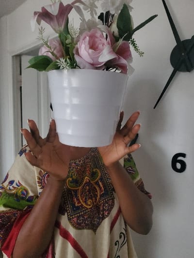 A person is positioned centrally, obscuring their face with a white, horizontally ribbed flower pot containing artificial flowers. The floral arrangement consists of pink peonies, pink and white lilies, and smaller white filler flowers with green foliage. The individual wears a loose-fitting garment patterned in cream, red, green, gold, and blue. Their dark-skinned hands, displaying manicured fingernails with white tips, are cupped around the lower sides of the pot. To the upper right, a minimalist black wall clock without a full face is mounted on a white wall, showing the numeral '6' and two black hands, indicating approximately 6:55. The background consists primarily of a plain white wall, with a partial doorway visible to the far left. The setting is indoors. No specific visual information in the image confirms the location as Festac Town, Nigeria.