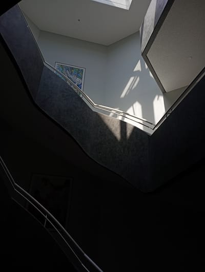 An interior shot of a modern staircase at an unknown location in Constance, Germany, features dramatic light and shadow play. Sunlight streams in from a skylight, casting geometric shadows across the white walls and grey, textured stairwell. A framed colorful artwork hangs on the upper wall. The staircase curves upwards, with dark, shadowed lower levels contrasting with the brightly lit upper sections. A white railing lines the stairs.