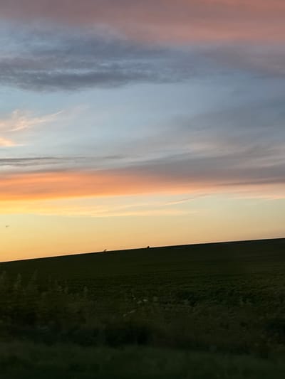 The image depicts a vast, open field under a colorful sky during sunset. The sky is a gradient of soft hues, transitioning from a pale yellow at the horizon to shades of orange, pink, and then blue and gray as it ascends. Streaks of clouds are visible across the sky, catching the warm light of the setting sun. The foreground consists of a dark, undulating field, likely agricultural land, with some vegetation and shadows creating texture. Two small, dark figures are discernible on the crest of the field, their forms silhouetted against the lighter sky. The overall impression is one of a serene, rural landscape at dusk. The scene is located in the city of Zvegor, Bulgaria.