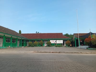 The image shows the exterior of a building complex, possibly a school, in Parung Panjang, Indonesia. The buildings are predominantly green with red-tiled roofs. The architecture appears simple and functional. A paved area made of interlocking blocks occupies the foreground, suggesting a courtyard or parking area. Signage is visible on the central building, partially obscured by bushes. A tall flagpole stands near the right-hand side of the image. The sky is clear and blue, indicating daytime. No people are visible in the scene.
