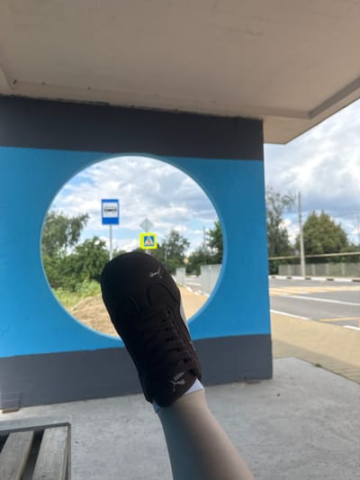In Shulhynka, Ukraine, a person is resting their leg, wearing a dark brown Puma sneaker with white accents and white socks, on a concrete surface next to a wooden bench. The shoe is positioned in the foreground, partially obscuring a circular blue cutout within a structure painted blue and gray. Beyond the cutout, a bus stop sign with a white bus icon on a blue square background is visible, along with a yellow pedestrian crossing sign. The background shows trees, a road with yellow markings, and a cloudy sky, suggesting an outdoor public area like a bus stop or transit shelter.