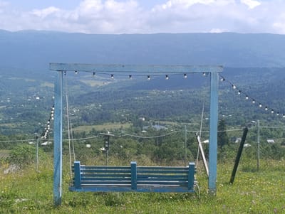 A blue wooden swing set stands on a grassy hilltop, overlooking a valley dotted with houses and trees. The swing set is adorned with a string of white globe lights that trail off to the right. In the background, layers of blue-tinged mountains rise against a cloudy sky. The scene appears to be in or near Nikortsminda, Georgia. The swing seat itself is constructed of horizontal blue planks. The surrounding area is overgrown with green grass and scattered small wildflowers. A wire fence runs horizontally across the mid-ground of the image, separating the swing area from the valley below.