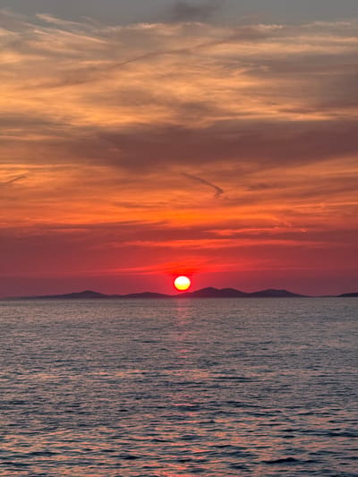 A vertical, medium shot captures a sunset over the Adriatic Sea, with a silhouetted archipelago in the background. The sky is ablaze with vibrant hues of orange, red, and pink, with wispy clouds streaking across the canvas. The sun, a fiery orb of yellow and red, is positioned low on the horizon, casting a warm glow over the scene. The sea is a deep blue, with gentle ripples reflecting the colors of the sunset. The islands, dark and jagged, create a dramatic contrast against the colorful sky. The image evokes a sense of tranquility and natural beauty, characteristic of a coastal sunset in Primošten, Croatia.