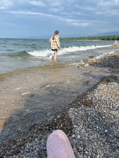 A person is walking in shallow water near a pebble beach. The person is wearing a plaid shirt and shorts. The sky is cloudy, and there are mountains in the distance. The water is calm with small waves breaking on the shore. The beach is covered in smooth, multi-colored pebbles. The foreground shows a pink object that appears to be a shoe or slipper. The scene is likely located at Lake Baikal near Tolbazikha, Russia.