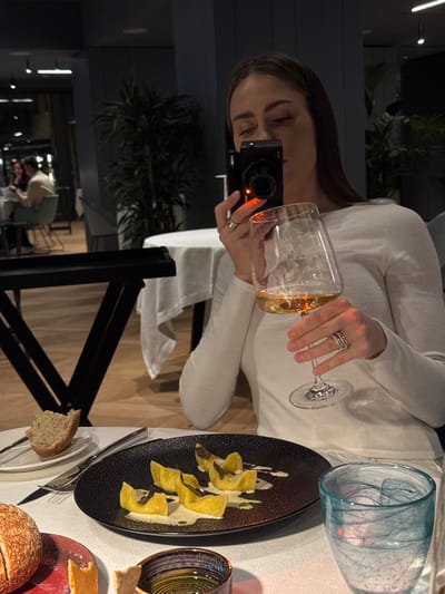 An upscale restaurant in Torbole, Italy, is depicted in a dimly lit indoor scene. In the foreground, a woman with long brown hair, wearing a white long-sleeved top, is seated at a table covered with a white tablecloth. She holds a black compact camera, exhibiting a red indicator light, to her face with her right hand, seemingly capturing a photograph. Her left hand holds a clear wine glass filled with light amber liquid. Multiple rings are visible on her fingers.
On the table in front of her, a black textured plate presents five pieces of yellow folded pasta, arranged on a light sauce and garnished with dark slices, likely truffle. To the left of the pasta plate, a small white plate holds a piece of white bread with a bite removed, alongside a knife and fork. A dark bread roll rests on a red-patterned plate. A small, dark-rimmed bowl contains a clear liquid, possibly olive oil, and a small food item. To the right, a clear glass with a distinctive light blue swirl pattern contains ice and water.
The background reveals a modern dining environment with dark blue walls, light wooden flooring, and several potted plants. Another white-clothed table is visible with two indistinct figures seated, appearing to be dining. A black serving trolley or stand is partially visible on the left side of the frame.