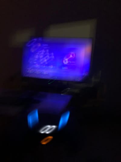 The image displays a dark, blurred indoor scene located in Acassuso, Argentina. The primary subject is a brightly lit, rectangular screen, likely a computer monitor or television, emitting a predominantly blue-purple light. The screen's content appears to be a graphical user interface; a distinct white-outlined icon, possibly a video game controller or a stylized guitar, is discernible on the right side against the blue background. Below the screen, a dark, flat surface, presumably a desk, is present. On this surface, a peripheral device is visible, characterized by two vertical, prominent blue light streaks and three horizontally elongated oval lights: two white and one orange, suggesting illuminated elements of a keyboard or gaming accessory. Green and white light trails, indicative of motion blur or light reflections, are present near the base of the screen. No individuals are visible, and no specific actions or interactions can be identified.