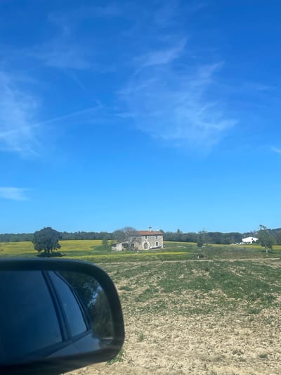 The image shows a rural landscape under a bright blue sky with some wispy clouds. In the distance, there is a building, possibly a farmhouse or villa. The vegetation consists of fields and some scattered trees. A side mirror of a vehicle is partially visible in the foreground, suggesting the image was taken from a car while in motion. The location is identified as Tordera, Spain.