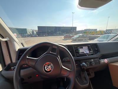 The image captures a first-person perspective from the driver's seat of a stationary Volkswagen commercial vehicle, positioned in an industrial area at Takkebijsters and Gieterijstraat in Breda, Netherlands. This location is confirmed by the active navigation system displayed on the vehicle's dashboard.
The foreground shows the vehicle's interior, including a black Volkswagen steering wheel prominently centered. To the left of the steering column, a red indicator light is illuminated, and a set of keys on a keyring is visible in a storage compartment. The dashboard's central infotainment screen displays a navigation map with a blue route line and road names such as "Takkebijsters" and "Gieterijstraat," along with "Breda." The windshield ahead is streaked and smudged, with visible glare from direct sunlight. A brown cardboard box is partially visible on the passenger side, and a white warning sticker depicting a crossed-out figure is affixed to the dashboard.
The exterior environment, viewed through the windshield, consists of a paved industrial or commercial parking lot under a clear, bright blue sky with direct overhead sunlight. In the mid-ground, a metal barrier separates the immediate vehicle area from the main lot. Multiple industrial buildings populate the background, including a prominent green-sided structure on the left and a darker, multi-sectioned building in the center-right. Several vehicles are parked in the lot, including a red hatchback, a silver hatchback, and a dark-colored sedan. Red and white traffic cones are scattered within the parking area, and tall light poles are visible. In the far right background, behind a fence, numerous yellow and green striped intermodal tank containers or industrial transport units are visible, indicative of a freight or logistics operation. No individuals are present in the scene.