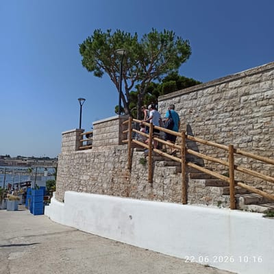 The image depicts a sunny outdoor scene in Trani, Italy, featuring a stone staircase leading upwards along a stone wall. A group of four individuals, two adults and two children, are ascending the stairs. The adults appear to be supervising or walking with the children. A wooden railing lines the staircase for support. At the top of the stairs, near the stone wall, is a tree with green foliage and a lamppost. In the background, a body of water, possibly the sea, is visible with some boats and structures along the shore. The foreground shows a concrete path and some blue crates. The date and time stamp "22.06.2026 10:16" is present in the lower right corner of the image. The architecture suggests a coastal Mediterranean setting.