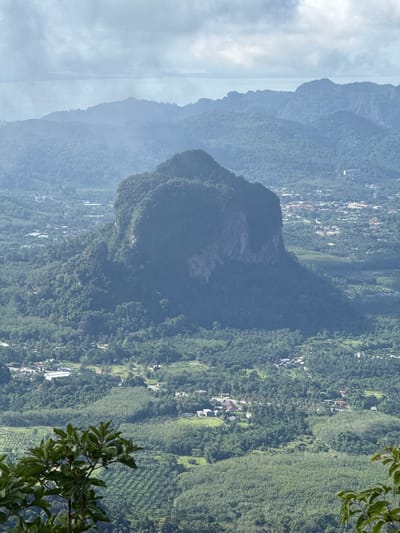 A high-angle, medium shot captures a dramatic mountainous landscape in Ban Din Daeng Nui, Thailand, under a hazy sky. In the foreground, the dark green leaves of a tree branch reach into the frame from the bottom left. The midground is dominated by a large, dark green, monolithic mountain with steep, rocky cliffs on its right side. This mountain is covered in dense vegetation.

Surrounding the central mountain is a sprawling vista of lush green hills and valleys. Small clusters of buildings, indicative of a town or village, are scattered throughout the landscape, some appearing to be residential. There are clearings and agricultural fields, likely rubber plantations given their characteristic orderly rows, visible among the dense forest. Roads wind through the terrain, connecting the scattered settlements.

In the background, receding layers of mountains fade into a soft, blue haze, and beyond them, a faint body of water is visible on the horizon, possibly the sea. The sky is cloudy, with patches of lighter and darker grey, suggesting a diffused daylight. There are no people or animals visible in the scene. The overall impression is one of natural grandeur and human habitation integrated within a verdant, tropical environment.