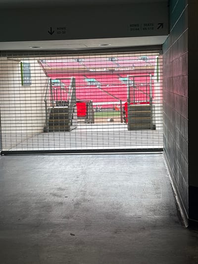 The image presents a view from within a dark, concrete tunnel or concourse, looking out into a brightly lit stadium through a black metal grating. The tunnel features grey concrete walls and a tiled ceiling.
Beyond the vertical metal grate, rows of vibrant red stadium seating are visible, indicating a large arena. On what appears to be the field level, several black structures with red accents are present, resembling stage equipment, large speakers, or temporary event installations. No people are visible, suggesting the image was captured before or after an event, or during the setup/teardown phase. The presence of stage-like equipment hints at a concert or a non-sporting event rather than a typical sports match.
On the left concrete wall of the tunnel, two white rectangular signs with black text are visible. The upper sign reads "ROWS | SEATS" with "37-44 | 95-110" below, accompanied by an arrow pointing right into the stadium. Further down, another sign indicates "ROWS" and "02-30" with an arrow pointing left. A small, partially obscured green sign is situated on the ground level near the bottom left of the tunnel opening, displaying "A&B" and what appears to be a time, possibly "08:56" or "09:56".
The natural light illuminating the stadium area suggests it is daytime. The scene is typical of a large event venue, consistent with the provided location context of Wembley, United Kingdom.