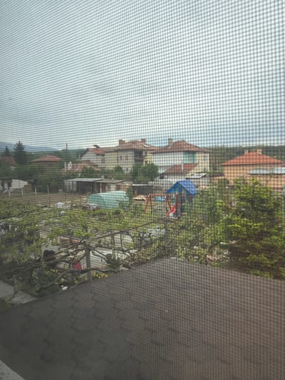 The image, taken from behind a screen, depicts a residential area in Barakovo, Bulgaria. Several houses with red-tiled roofs are visible in the background. A children's playground with blue and red play structures is located in the foreground amongst trees and greenery. There are also some small sheds and tents among the vegetation. The image has an obstructed view due to the screen mesh.