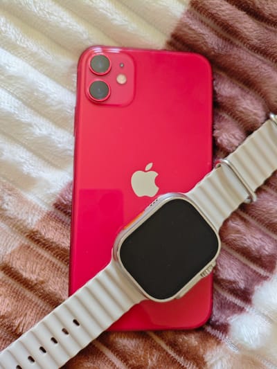 This image depicts a close-up, overhead view of a red iPhone and a white Apple Watch with a textured strap. The iPhone is positioned vertically, with its camera bump visible at the top left, featuring two lenses. The Apple logo is centered on the back of the iPhone. The Apple Watch is placed diagonally across the iPhone, with its screen facing up and its strap extending towards the bottom left of the frame. Both devices are resting on a surface covered with a textured fabric, appearing to be a blanket or throw, with alternating bands of light beige and pinkish-brown. The overall lighting suggests an indoor setting, with soft illumination. There are no people visible in the image. The scene does not contain any recognizable landmarks or features that would indicate a specific location within Sumgayit, Azerbaijan, or any other city.