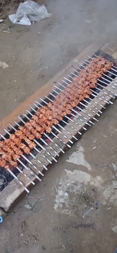 In Daliguzar, Uzbekistan, a barbecue is underway with numerous skewers of raw, seasoned meat cooking over hot coals. The skewers are arranged in rows on a makeshift grill constructed from bricks and metal rods. Smoke rises from the grilling meat, indicating the cooking process. The surrounding environment appears to be an outdoor area with a dirt or gravel ground, and some scattered debris including a white plastic bag. The scene depicts a casual outdoor cooking event.
