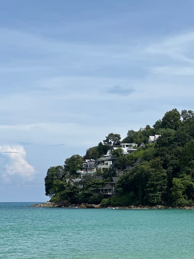 The image displays a coastal scene in Ban Bang Thao, Thailand, featuring a lush green hillside dotted with white buildings and a turquoise body of water in the foreground. The hillside slopes down towards the sea, with the buildings strategically placed among the dense foliage, some appearing to be villas or resort accommodations with dark tiled roofs. The water is calm with subtle ripples, and on the left side of the frame, a large cumulus cloud formation is visible against a pale blue sky with scattered cirrus clouds. The rocky shoreline is visible where the land meets the sea. No people are discernible in the image.