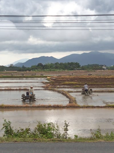 The image depicts a wide view of flooded rice paddies in Diên Lạc, Vietnam. The sky is overcast with dark, dramatic clouds, and a range of mountains forms the backdrop. Several individuals are operating small tillers in the shallow water of the paddies, preparing the land. One person, wearing a hat and a light-colored shirt, is clearly visible in the foreground, working a tiller. In the middle ground, another person is also seen operating a tiller, and further back, additional figures and their machinery are discernible. The paddies are divided by earthen banks, and the water reflects the muted light. Vegetation lines the edges of the paddies and the foreground, leading to a paved road at the bottom of the frame. Power lines are strung across the top of the image.