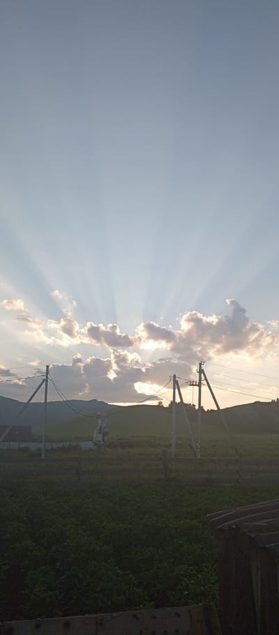 In the city of Khusainovo, Russia, a rural landscape is depicted at dusk. The sky is a pale blue, transitioning to a warmer hue near the horizon, where the sun is setting behind rolling hills. Beams of sunlight radiate upwards from behind scattered, fluffy clouds, creating a dramatic crepuscular effect. In the foreground, a field of dark green vegetation stretches across the lower portion of the frame. A wooden fence runs along the middle ground, separating the field from what appears to be a power substation with utility poles and wires. In the far left, a portion of a dark-roofed building is visible. The overall scene conveys a sense of peaceful, rural twilight.