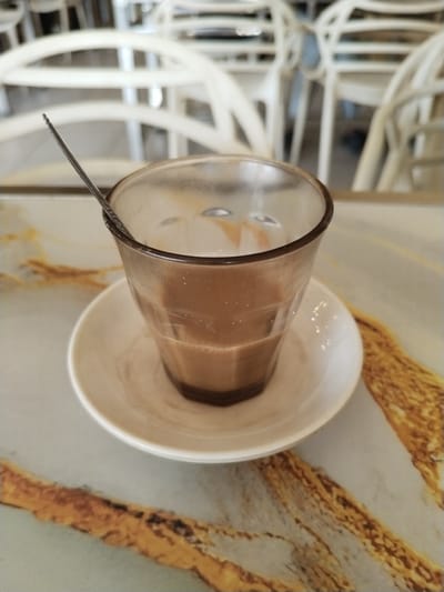 A slightly used glass of coffee rests on a saucer on a table. A spoon is in the glass. The table has a marbled surface, and white chairs are visible in the background. The setting appears to be a cafe or restaurant. There are no people present in the image. Location unspecified.