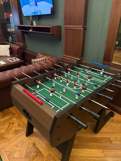 A foosball table is the central focus of the image, positioned on a wooden parquet floor. The table itself is dark brown with a green playing surface and white markings. It is populated with miniature football players in red and blue uniforms, arranged on metal rods. The table has black handles for control.

To the left of the foosball table, a plush, brown leather sofa is visible, with a white cushion and a magazine on a coffee table in front of it. Behind the sofa, a dark wooden shelf is mounted on a teal-colored wall. A television screen displaying a sports game is mounted above the shelf. To the right of the foosball table, a section of a dark wooden cabinet and a partial view of a mirror with a wooden frame are present. The overall environment appears to be an indoor recreational space, possibly a lounge area.

The image is set in Zhukovka, Russia, in a location that suggests a private residence or a club. The lighting is moderate, casting some shadows and highlighting the textures of the furniture and flooring.