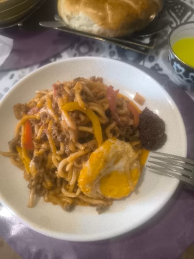 This image depicts a close-up, elevated view of a dining table setting in Turkkishlak, Uzbekistan. The central subject is a white, oval-shaped plate holding a meal composed of thick, light-brown noodles mixed with ground meat, strips of yellow and red bell peppers, and a portion of a fried egg or omelet featuring a distinct yellow yolk and white. A small, dark, granular condiment is present on the right side of the plate. A silver four-tined fork rests on the lower right edge of the plate. In the blurred background, a large, round, light-colored bread loaf, likely traditional Uzbek non, is visible on a dark tray. To the right of the bread, a small, white bowl with dark blue decorative patterns contains a yellow liquid. The table is covered by a patterned tablecloth displaying purple and lighter-toned abstract designs. No individuals or direct actions are visible.