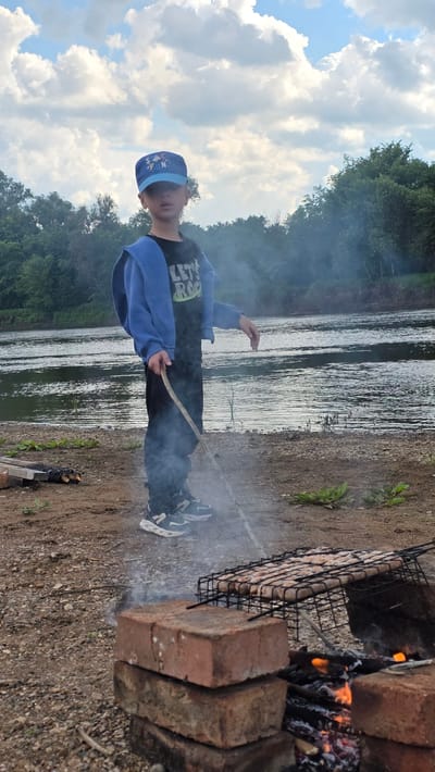A young boy wearing a blue baseball cap and a blue hooded jacket stands on a sandy bank next to a river. He is holding a stick. In front of him, a grill with food is cooking over a fire built with bricks. The sky is cloudy and the surrounding area is covered with trees. This scene appears to be in the city of Дарьино, Russia.