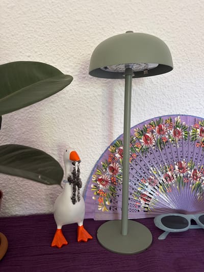 The image shows a collection of decorative items mounted on a textured white wall and a dark purple surface. A green lamp with a circular base and an angled arm extends from the wall. Attached to the arm is a decorative purple fan with a floral pattern in red, pink, green, and yellow. To the right of the fan, a pair of light blue sunglasses with dark lenses are perched vertically. Below the fan, a white decorative goose figurine lies on its side, with orange feet and a black, ornate decoration on its back. Two dark green leaves from a plant are visible in the lower left and lower middle of the image. The scene appears to be indoors, possibly a wall display in a home or shop in Las Rozas de Madrid, Spain. No specific time of day or weather conditions can be determined. There is no visible text in the image.