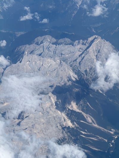This aerial photograph captures a vast mountain range with rugged, rocky terrain. The mountains are predominantly gray, with patches of snow and ice visible on some of the higher peaks. The lower slopes are covered in dark green vegetation. Several valleys and ravines carve through the mountains, with a river or stream visible in one of them. Clouds partially obscure some areas of the landscape, casting shadows and adding depth to the scene. In the distance, a small town or village with scattered buildings can be seen nestled in a valley. The overall impression is one of immense scale and natural beauty. The image does not depict the city of Leno, Italy; rather, it showcases a mountainous region.