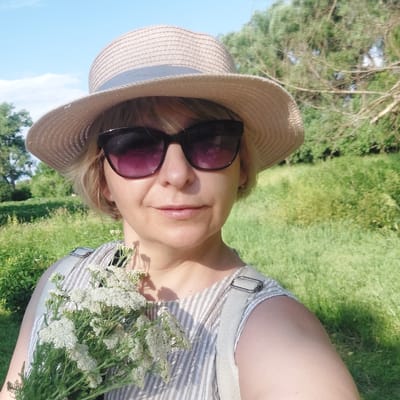 A woman is taking a selfie in an outdoor environment. She is wearing a straw hat with a light grey ribbon and large sunglasses with purple lenses. She has short blonde hair and is holding a bouquet of small white flowers with green stems in her left arm. She is also wearing a light-colored striped dress with light grey straps visible over her shoulders. The background consists of green grass and trees under a clear blue sky. The location is identified as the village of Voronkiv, Ukraine.