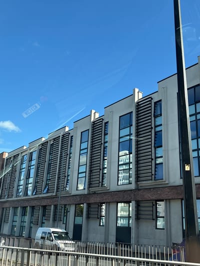 The image shows a modern building with multiple floors and rectangular windows, each featuring horizontal slats over them, taken from a low angle, looking upwards. The building's facade alternates between grey concrete and brick sections. A white van is visible at the bottom right of the frame, partially obscured by a grey metal railing. The sky above is bright blue with a few faint clouds. The image is taken from inside a vehicle, as evidenced by the reflection of a registration plate (KS3 COA) on the window. The building is a modern structure with distinct architectural design elements. This image was taken in Chester, United Kingdom.