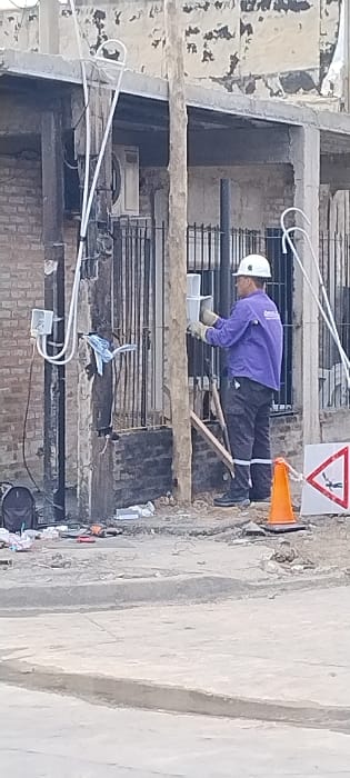 The image depicts a construction worker engaged in a task within an unfinished multi-story building. The worker, a man, is dressed in a white hard hat, a purple long-sleeved shirt with a logo on the back, black work pants with reflective bands near the ankles, and light-colored work gloves. He is bent over, appearing to be working with a white container or bucket positioned on a concrete beam, possibly mixing or applying materials.

The immediate environment is a construction site, characterized by exposed concrete beams, rebar extending from the structure, and visible brickwork on the walls. Below the worker's level, an air conditioning unit or similar appliance is partially visible amidst further exposed rebar and debris. White conduit pipes and electrical wiring are present, with a small white electrical box attached to a lower beam. A wooden plank lies near the worker's feet.

To the right of the worker, an orange traffic cone is positioned. Above and slightly to the right of the cone, a white triangular safety sign with a red border and a black pictogram of a falling person indicates a hazard, likely signifying a "falling hazard" or "slippery surface" warning. The background reveals textured concrete walls and raw building elements. The scene is brightly lit, suggesting it is daytime. The overall impression is an active, dusty construction site in San Fernando, Argentina.