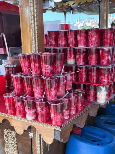 The image shows a street food stall in Fatih, Istanbul, Turkey. The stall is displaying many plastic cups filled with a pinkish-red liquid and shredded vegetables, likely pickled turnips, a common Turkish street food. The cups are arranged on wooden shelves. Behind the cups, a large glass jar filled with the same red liquid is visible. A red ladle rests on the edge of the jar. The stall itself has a rustic wooden structure with some decorative carvings. In the background, a cityscape with buildings and a bridge can be seen under a bright blue sky, suggesting an outdoor location. Blue plastic containers are stacked to the right of the stall.