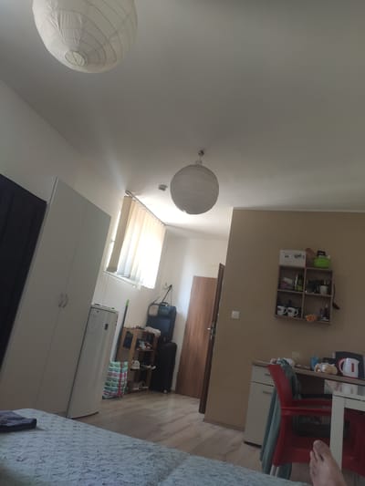 The image shows a bedroom in a residential setting, possibly in Tatabánya, Hungary. The scene is captured from a high angle, looking down into the room.
Prominent objects in the room include two large white paper lanterns hanging from the ceiling, one in the foreground and another further back. To the right, a white shelving unit is mounted on a light brown wall, containing various items such as bottles, boxes, and decorative mugs. Below the shelves, a red chair and a white desk with a red chair are visible. A white filing cabinet is beneath the desk.
Further into the room, a closed white wardrobe with two handles is present. Next to it, a window with light-colored blinds is letting in natural light. Adjacent to the wardrobe, a white refrigerator or washing machine stands. Behind this appliance, a dark blue suitcase and a black bag are stacked on the floor. A small wooden shelf unit is positioned between the suitcases and the appliance, holding several items. The flooring is light wood.
A person's foot, with toes visible, is in the top right corner of the image, suggesting a person is present in the room but not fully captured.
The lighting suggests it is daytime, with natural light coming through the blinds. The overall atmosphere appears to be that of a lived-in bedroom. No specific activity or event is clearly taking place, other than the implied presence of someone inhabiting the space. There is no visible text in the image.