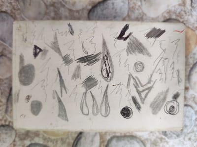 The image shows a close-up, overhead view of a rectangular piece of paper covered in abstract pencil and crayon scribbles. The paper appears to be old and slightly worn, with some discoloration. The doodles include various shapes and lines, such as zigzags, triangles, circles, and shaded areas. There are also some irregular blobs and what appear to be stylized teardrop shapes. The paper is placed on a surface composed of smooth, rounded stones of various sizes and colors, predominantly grays, whites, and beiges. The lighting suggests an indoor environment. There is no indication of the location being in Termal'nyy, Russia, or any specific city.