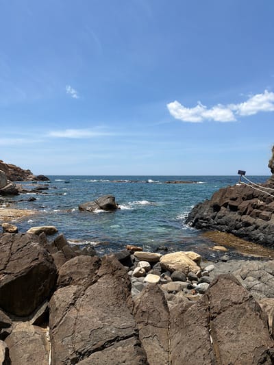 The image shows a rocky coastline on a clear day. The ocean is a deep blue with waves breaking against the shore. There are several large rocks in the foreground and mid-ground, some of which are partially submerged in the water. In the distance, the ocean stretches to the horizon, meeting a bright blue sky with a few white clouds. A small signpost is visible on a rock ledge on the right side of the image, with white ropes leading away from it. The scene appears to be a natural coastal landscape, possibly a public area given the signpost and ropes, suggesting it might be a designated viewpoint or a safe access point for visitors. The general impression is one of a tranquil and rugged natural environment. The location is identified as Phú Lương, Vietnam, a coastal region known for its scenic beauty.