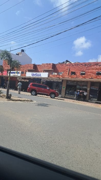 The image presents a daytime street scene in Majengo ward, Kenya, viewed from a slightly elevated position, possibly from a vehicle. The sky is clear blue with sparse white cumulus clouds, crisscrossed by numerous overhead power or communication lines.

An asphalt road dominates the lower half and foreground of the image. On the right side, a row of two-story commercial buildings runs parallel to the street. The upper story of these buildings features exposed reddish-brown brickwork. Ground-level storefronts include various shops, with a prominent white sign displaying "UMOJA" in blue lettering centrally located above one entrance. A smaller, illegible sign is visible to its left.

A red SUV, appearing to be a Toyota Land Cruiser Prado or similar model, is parked curbside in front of the buildings, angled slightly towards the right side of the frame. One male subject, dressed in a light-colored shirt and trousers, is walking on the sidewalk towards the right, positioned near the front of the red SUV. Further along the sidewalk to the right, a group of approximately three to five individuals stands near a storefront and a parked motorcycle.

To the left of the red SUV, a small palm tree stands on the sidewalk. Behind the commercial buildings and to the left, a taller white building with a dark water tank on its roof is visible. Strong sunlight creates distinct shadows across the road and buildings, with a large shadow from an unseen object in the immediate foreground covering the bottom-left portion of the asphalt.