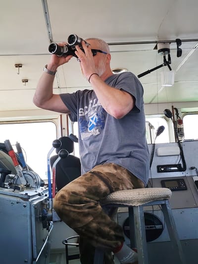 A middle-aged man is seated on a stool in the bridge of a vessel, possibly in Koprino, Russia. He is wearing a grey t-shirt with a blue logo and text that appears to say "Scotland," camouflage pants, and sunglasses. He is holding a pair of binoculars up to his eyes and looking forward. He has short grey hair and a grey beard. A silver wristwatch is visible on his left wrist. The interior of the bridge has various controls and equipment, including levers, gauges, microphones, a radio, and a white bucket hanging from the ceiling. The controls are painted a utilitarian grey. Red and blue are visible on the control levers. A window is in the background, suggesting the man is likely looking out at the surrounding area.