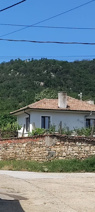 The image shows a building with a red tile roof and white walls. The building has a chimney, windows, and a stone wall along its side. A green, tree-covered hillside is visible in the background. Overhead power lines are visible in the sky. A concrete pavement is visible on the right. The location is Beguntsi, Bulgaria. No people are visible.