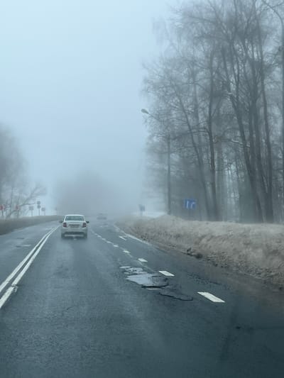 The scene depicts a multi-lane asphalt road in Butyrki, Russia, on a day characterized by dense fog and limited visibility. The road surface is wet and exhibits several potholes, some containing standing water. Lane markings include a double solid white line on the left and broken white lines on the right, delineating lanes.
A white sedan with illuminated taillights is in the foreground, traveling away from the viewer. Its rear license plate appears to be Russian, partially legible as "P796XM 197". Two additional vehicles are faintly visible further ahead on the road, receding into the fog.
To the right of the road, a bank of dirty snow or slush borders the pavement. A line of bare deciduous trees stands further back, partially obscured by the fog. A streetlight on a pole is visible among these trees, and a blue road sign with white arrows indicating a straight path and a right turn is present. Another similar sign is faintly visible further down the road.
The left side of the road shows bare trees and what appear to be utility poles or structures extending into the fog. Distant red and white striped warning barriers or signs are barely discernible through the heavy mist. The sky is uniformly gray and overcast, contributing to the overall hazy and muted light conditions. No people are visible.