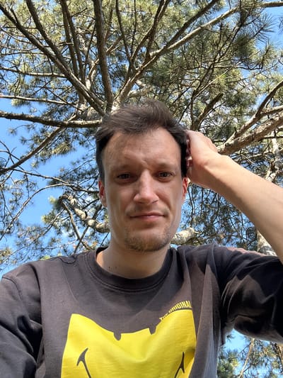 A man with short, dark hair and stubble is looking directly at the camera, his right arm raised and resting on his head. He is wearing a dark t-shirt with a large yellow smiley face graphic and some text that reads "THE ORIGINAL". The background is dominated by the branches and needles of a pine tree against a clear blue sky. The image appears to be a selfie taken outdoors, possibly in Velykozalissia, Ukraine, given the context.