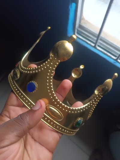 The image presents a close-up view of a person's hand holding a golden crown. The hand has a dark skin tone, with fingers gently gripping the base of the crown. The crown itself is a shiny, bright gold, likely made of plastic or a light metal. It features a traditional design with multiple upward-curving points, each topped with a small, rounded finial. The surface of the crown is decorated with rows of small, raised bumps. Embedded into the crown are several simulated gemstones; visible ones include a circular blue stone and an oval green stone, enhancing its regal appearance.

The scene takes place indoors, with a dark, blurred background. Natural light filters in from the upper right, highlighting a window structure that appears to have a grid pattern, possibly bars, and a prominent blue frame. The lighting suggests it is daytime. No specific activity beyond holding the crown is evident. There is no visible text in the image. This scene is located in Warri South, Nigeria.