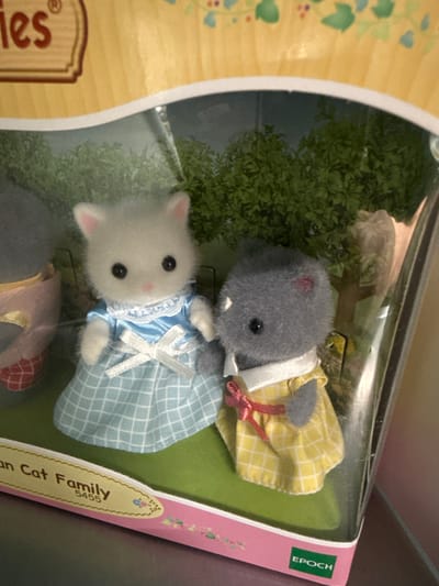 The media file shows two plush toy cats inside a clear plastic toy box. One cat is grey and wearing a yellow dress with white trim and a red ribbon. The other cat is white and wearing a blue checkered dress with white lace trim and a white bow. Both cats have black eyes and pink accents on their ears and cheeks. The background inside the box appears to be a green, leafy artificial landscape. The toy box is partially visible, showing a pink and yellow cardboard backing with floral illustrations and text. Visible text on the backing includes "EPOCH", "an Cat Family", and "5455". The setting is likely a retail environment given the toy packaging. The image is taken from a slightly elevated angle, with the toys oriented vertically within the frame.
