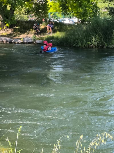 The image depicts two individuals in a blue inflatable raft, navigating a flowing river. Both individuals are wearing red helmets and life vests. The water is green, suggesting the presence of algae or sediment. On the riverbank, there are trees and grass. Two more individuals are visible on the riverbank. Based on the location, it can be stated that the scene is Saint-Pierre-de-Bœuf, France.