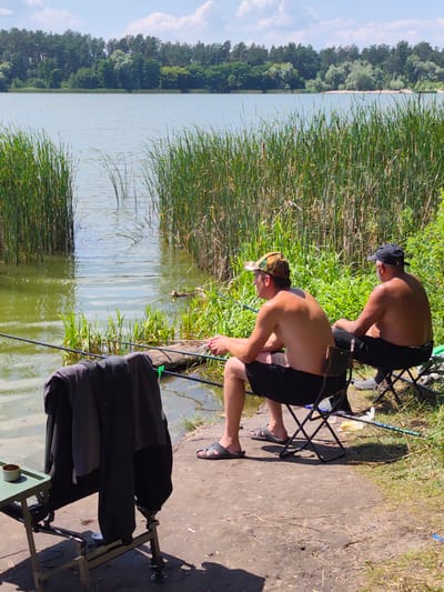 Two men are fishing on the bank of a lake, surrounded by tall reeds and greenery. The man on the left is wearing a camouflage cap and black shorts, while the man on the right has a black baseball cap and black pants. They are both sitting on folding chairs, with fishing rods extended over the water. A small green table with a cup sits next to the man on the left, and a dark jacket is draped over a fishing rod support. In the background, a dense forest lines the opposite bank of the lake. The water is calm with reflections of the sky and trees. The scene appears to be a peaceful, natural setting, likely within Bobrytsia, Ukraine, given the context provided.