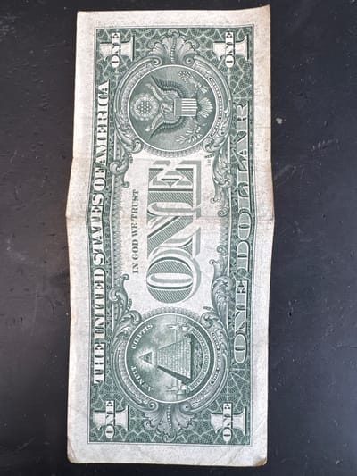 The image shows the reverse side of a U.S. one-dollar bill. The bill is oriented vertically, with the top edge at the top of the image. It is lying on a dark, textured surface, possibly a table or countertop. The bill itself is creased and slightly worn, with visible folds.
The design elements on the reverse of the bill are clearly visible. At the top center is the Great Seal of the United States, featuring an eagle with outstretched wings holding an olive branch and arrows. Above the eagle is a banner with the Latin phrase "E PLURIBUS UNUM." Below the eagle is a pyramid with an eye at its apex, surrounded by the Latin phrases "ANNUIT COEPTIS" and "NOVUS ORDO SECLORUM." Large numerals and the word "ONE" are present in the corners and center of the bill. The words "THE UNITED STATES OF AMERICA" are printed vertically along the left side, and "ONE DOLLAR" is printed vertically along the right side.
There is no indication of the city of Mejaniga, Italy, in the image. The image depicts only the dollar bill and its background.