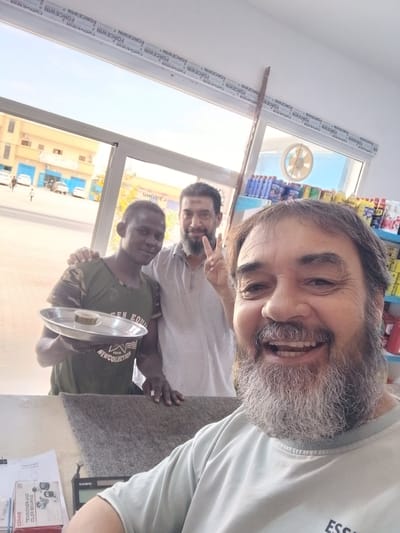 In Hun, Libya, three men are photographed indoors, possibly in a shop. A man in the foreground, with a grey and white beard, smiles broadly at the camera, his mouth open. Behind him, slightly to the left, a second man with dark hair and a beard makes a peace sign with his fingers. He is wearing a light grey shirt. To the left of the second man, a third man, who appears younger and has dark skin, holds a silver tray with a small cup on it. He is wearing a camouflage-patterned t-shirt. In the background, shelves are visible, stocked with various items, and a fan is mounted on the wall. Outside the window, buildings and cars can be seen. The overall lighting suggests daytime.