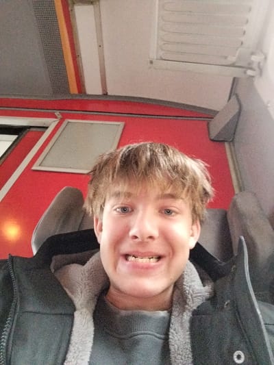 A young man smiles broadly, showcasing his teeth. He is seated, possibly on a train, given the red and grey interior with visible window frames and overhead compartments. He wears a dark jacket with a fleece collar over a grey sweater. His hair is blonde and tousled. The lighting appears to be natural, with a slightly grainy quality to the image. The angle is a selfie, taken from slightly below his face.